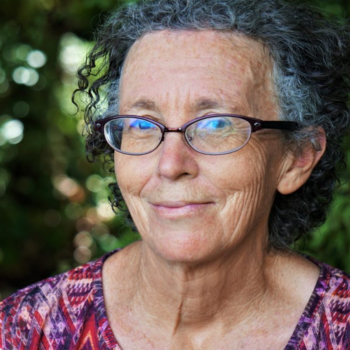 Smiling older woman with curly grey hair and glasses, wearing a patterned top, photographed outdoors with greenery in the background.